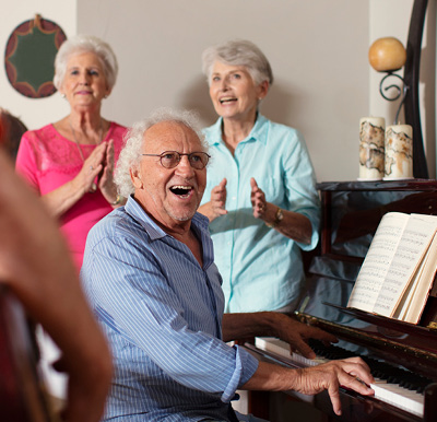 Senior man playing piano with friends gathered and clapping at senior living community