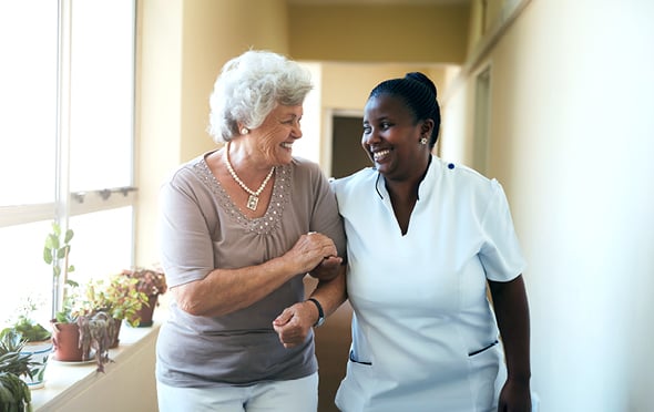 Smiling caregiver assisting a resident down a hall with large window and potted plants.