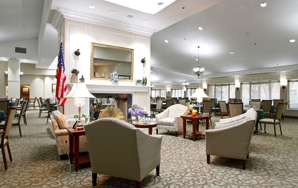 Large, elegant dining room with multiple tables, a fireplace in the center and lounge seating at Olde Raleigh assisted living community in Raleigh, NC.