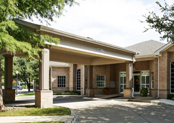 The front entrance of an assisted living community in Richardson Texas, with a portico, large trees and potted plants.