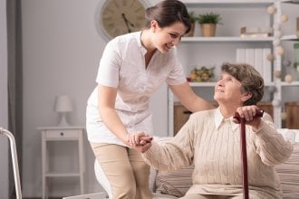 Caregiver helping a senior woman with a cane sit down.