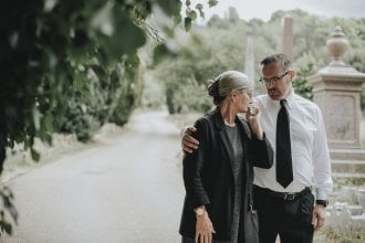 Senior woman dressed in black is embraced by younger man after a funeral.