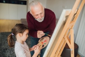 Senior man helps young girl paint on an easel
