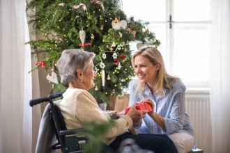 Senior woman receives gift from younger woman in front of Christmas tree.