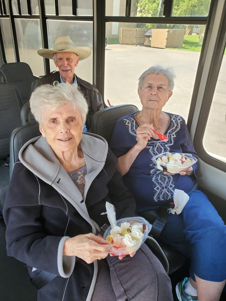 two senior women and a senior man smile while eating banana splits in the community car