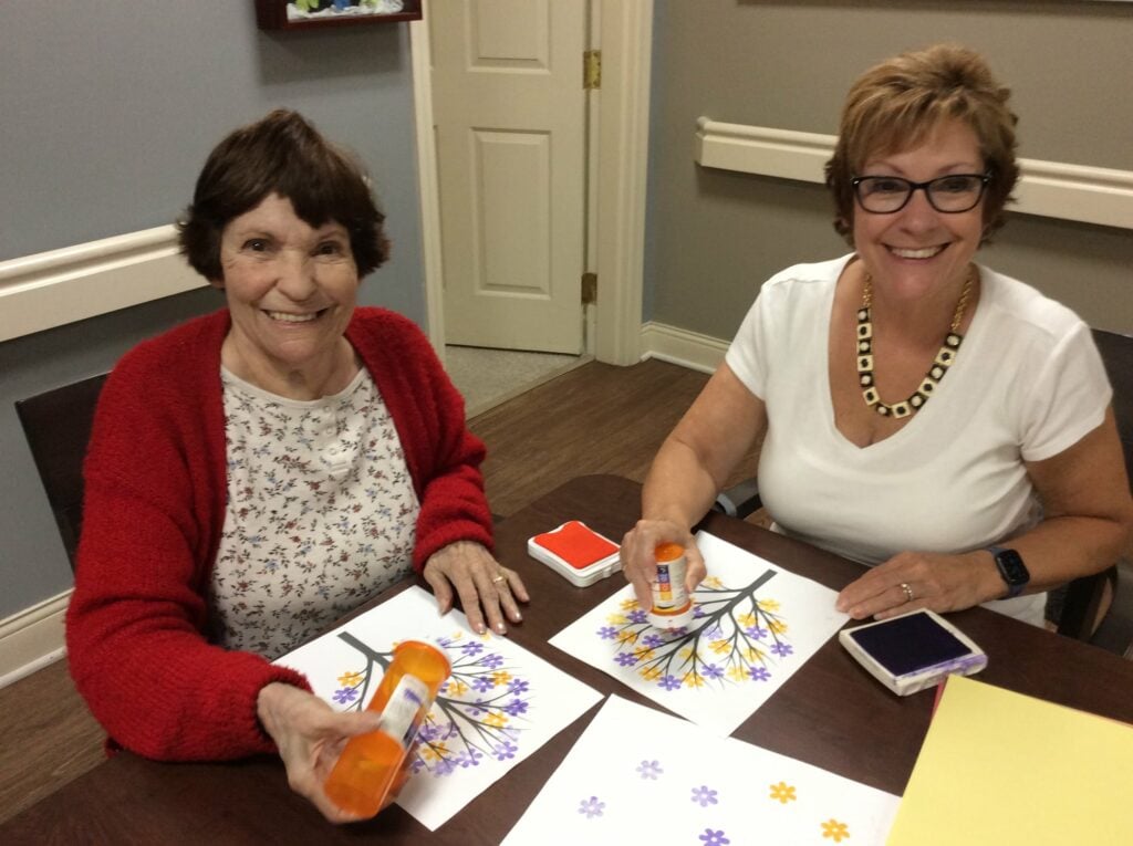 two senior women do a painting craft together and smile at the camera