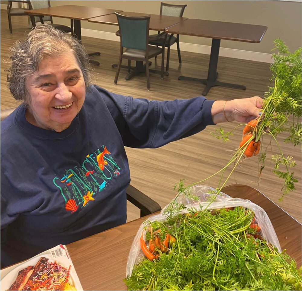 Senior woman smiles while holding a carrot in her hand at a senior living community in Lambertville, Michigan