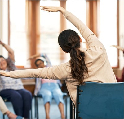 Caregiver leading seniors in a chair yoga stretching exercise.