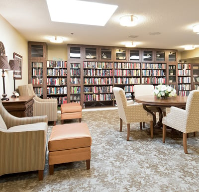 Beautiful library in senior living facility with built-in wood shelving and cabinetry along the back wall, comfortable arm chairs and a table in the center of the room in Omaha, Nebraska.