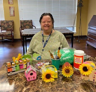 A resident smiling behind a table filled with craft supplies and decorated bird houses in Fort Worth, Texas.