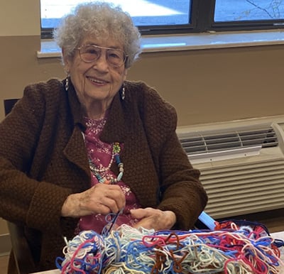 senior woman smiles while she knits at a table
