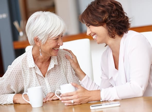 A caregiver smiling and putting her hand on a resident's shoulder while they enjoy coffee together.