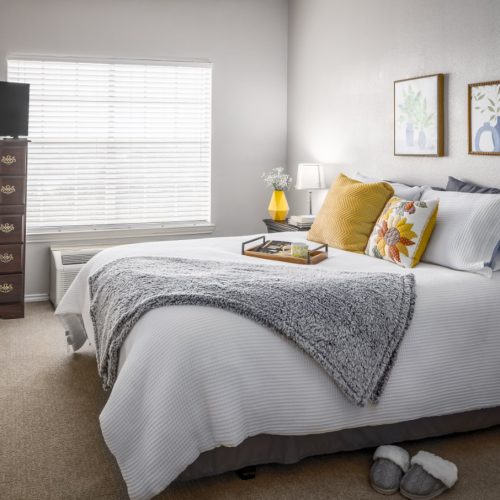 Apartment bedroom includes a large bed, side table, window, dresser and a small TV
