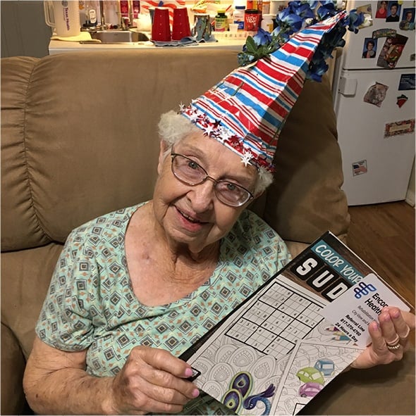 A smiling resident wearing a silly hat and holding a sudoku, coloring book in her apartment in North Richland Hills, Texas.