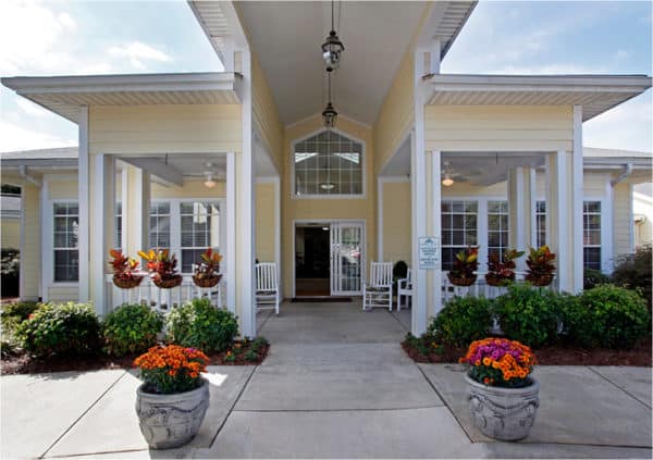 Front entrance of a senior living facility in Anderson, South Carolina with colorful flowers lining walking and white rocking chairs at entrance.