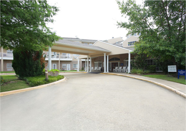 Gray senior living community in Mesquite, Texas with a long driveway and rocking chairs on the front porch and a retirement community sign.