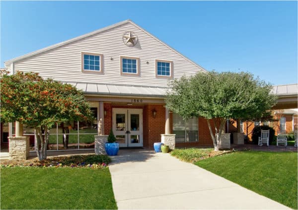 The entrance to an assisted living community with sidewalk, flowers and white rocking chairs in Arlington, Texas.