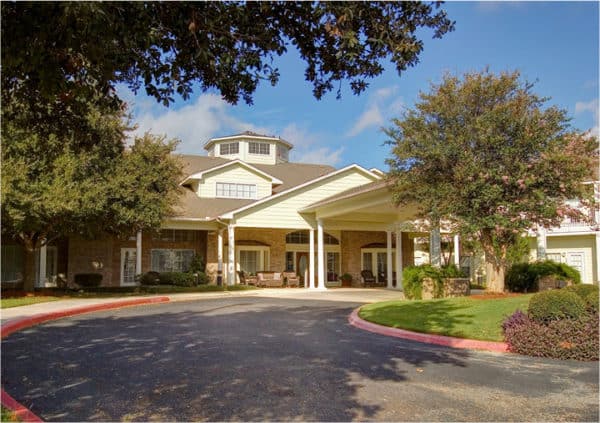 The exterior of a senior living community in San Antonio, Texas, with comfortable seating on a covered patio, large trees and beautiful landscaping.