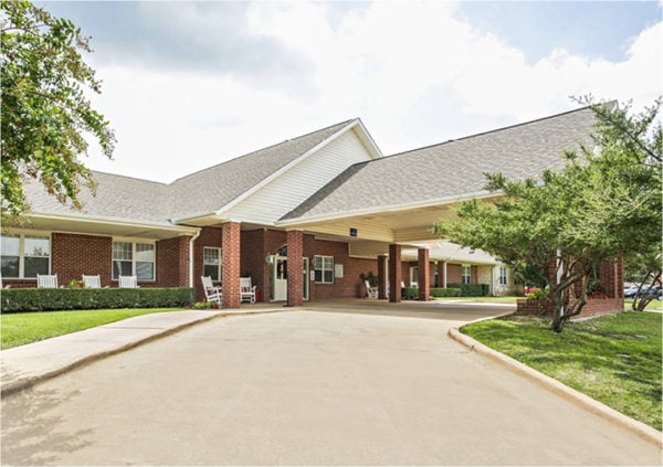 Front entrance of a senior living facility in Keller, Texas with covered walkway and white rocking chairs.