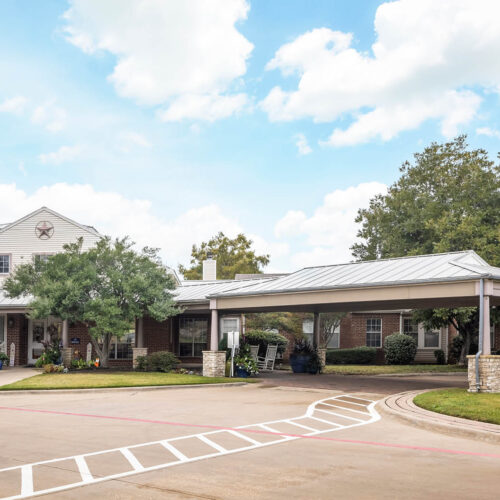 The Waterford on Cooper - Senior living in Arlington, Texas. The entrance to an assisted living community with sidewalk, flowers and white rocking chairs in Arlington, Texas.