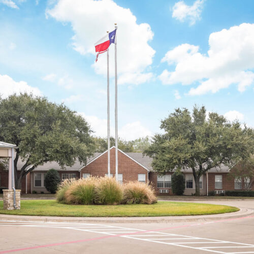 The Waterford on Cooper - Senior living in Arlington, Texas. The entrance to an assisted living community with sidewalk, flowers and white rocking chairs in Arlington, Texas.