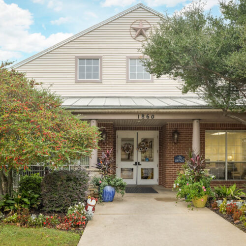 The Waterford on Cooper - Senior living in Arlington, Texas. The entrance to an assisted living community with sidewalk, flowers and white rocking chairs in Arlington, Texas.