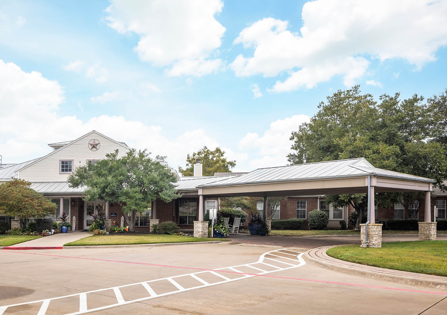 The Waterford on Cooper - Senior living in Arlington, Texas. The entrance to an assisted living community with sidewalk, flowers and white rocking chairs in Arlington, Texas.