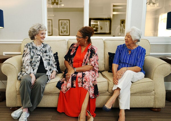 Three older adult females sit together on a couch while smiling and laughing at a senior living community