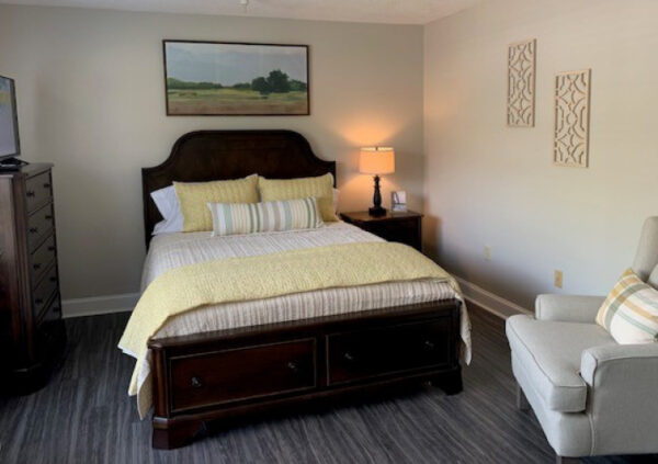 Assisted living apartment bedroom with a large bed, chair, dresser, TV and night stand