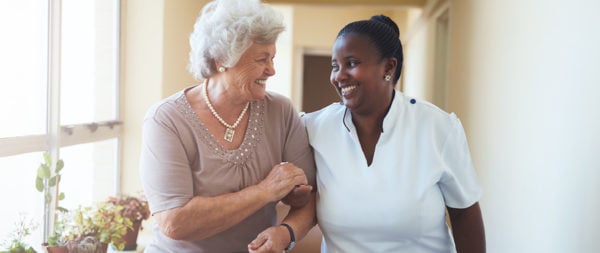 Senior woman being led down the hall by a smiling caregiver.