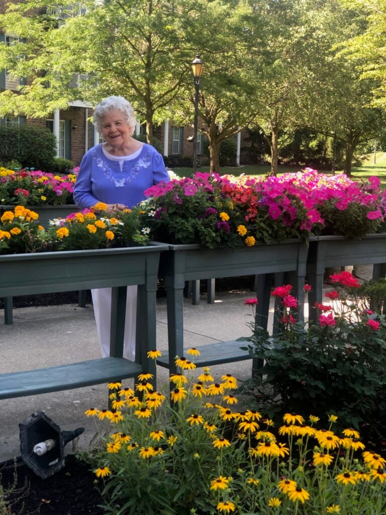 Senior woman smiles while standing among flowers at a senior living community in Fort Wayne, Indiana.
