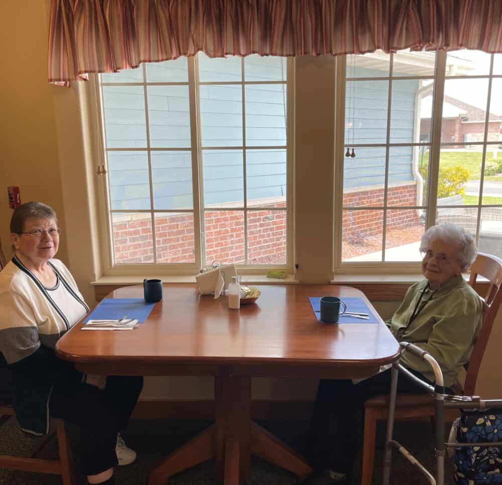 Two senior women smile at the camera while getting ready to eat at a senior living facility in Colby, Wisconsin.