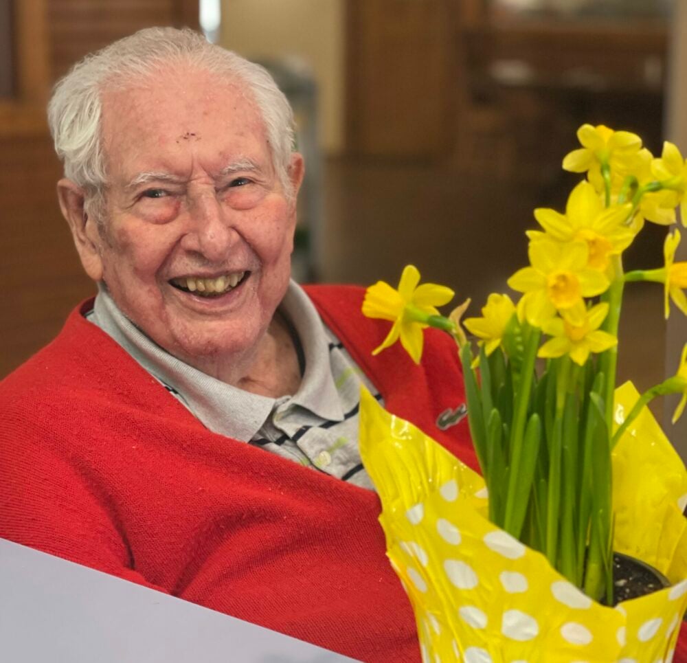 senior man smiles near a bouquet of yellow flowers
