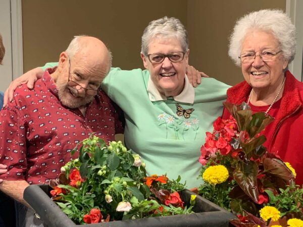 two senior women and a senior man smile while planting flowers together