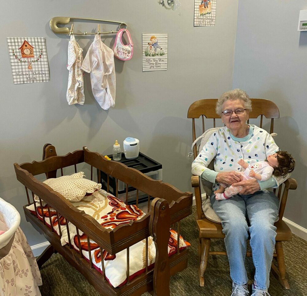 senior woman smiles while holding a baby doll near a crib