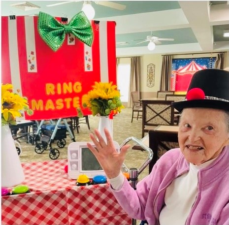 Senior woman smiles and waves in front of a ring master display at a senior living community in Indianapolis, Indiana.