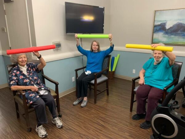 three senior women do exercises together by lifting pool noodles above their heads