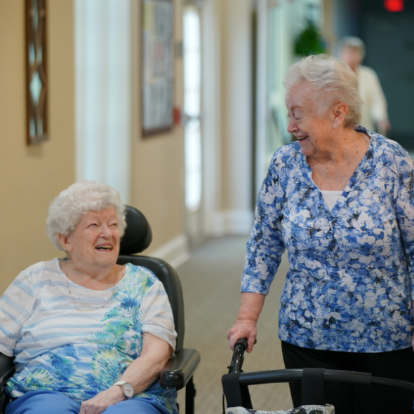 Two senior women smile and laugh while walking in a hallway