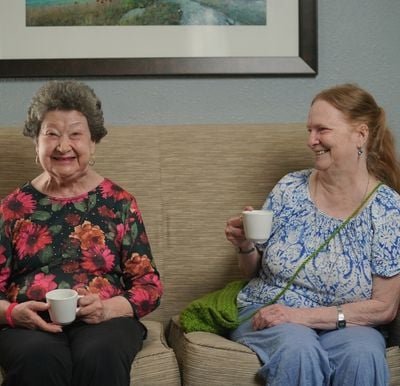 two senior women sitting a couch together with a drink in hand while laughing