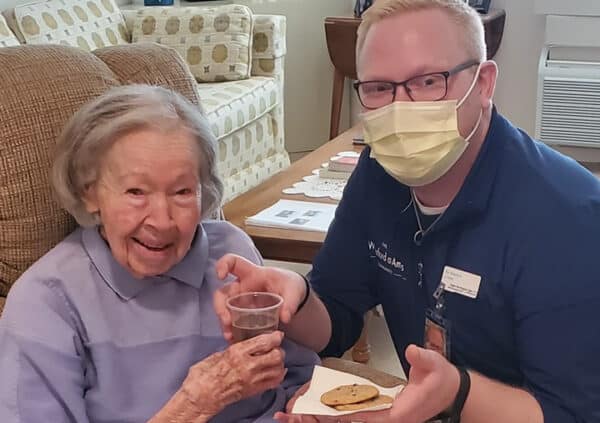 A caregiver wearing a mask and serving chocolate chip cookies and a beverage to a smiling resident.