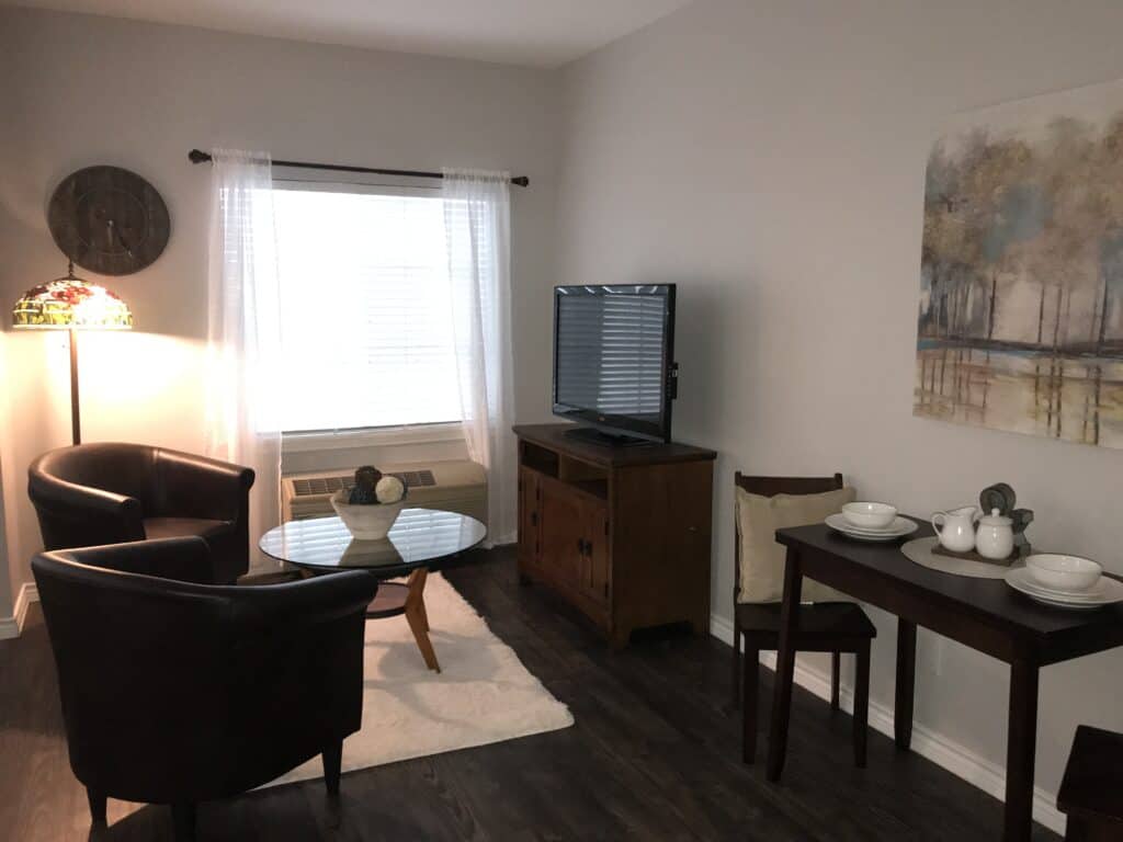 Studio apartment with lounge area, tv, chairs and near window at a senior living facility in Corpus Christi, Texas.