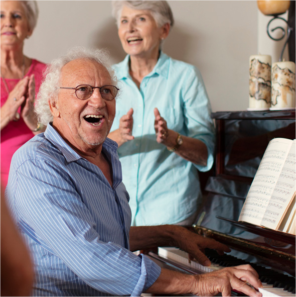 A senior smiling and playing piano while fellow residents sing and clap along in their senior living retirement community.