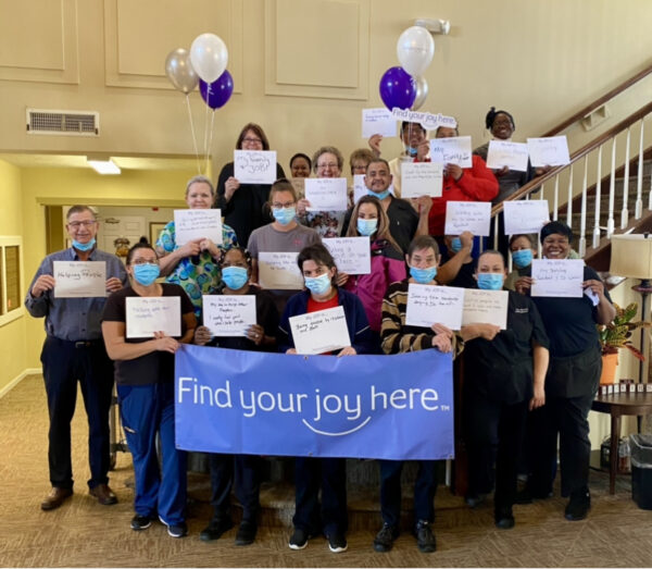 large group of employees hold signs that say "my joy is..." in front of a find your joy here sign
