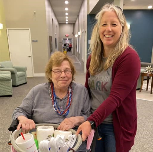 woman helps senior woman smile and pose for the camera while holding a bin of supplies