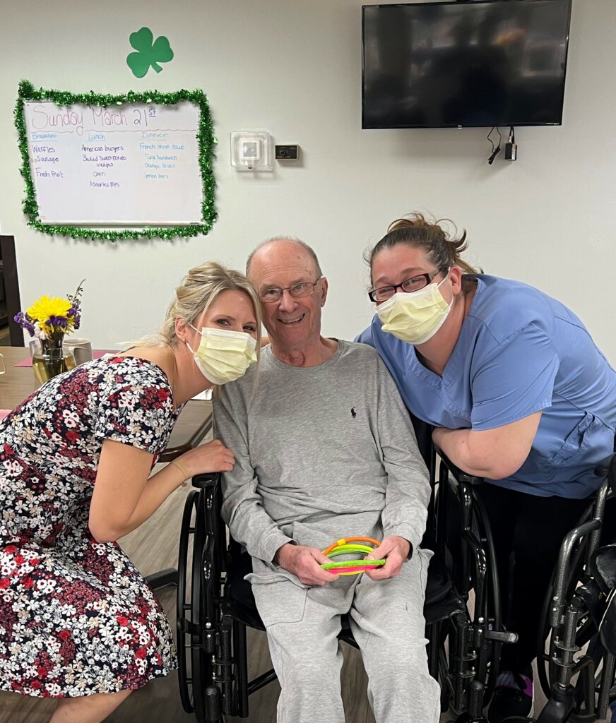 two women caregivers hug a senior man resident in his wheelchair