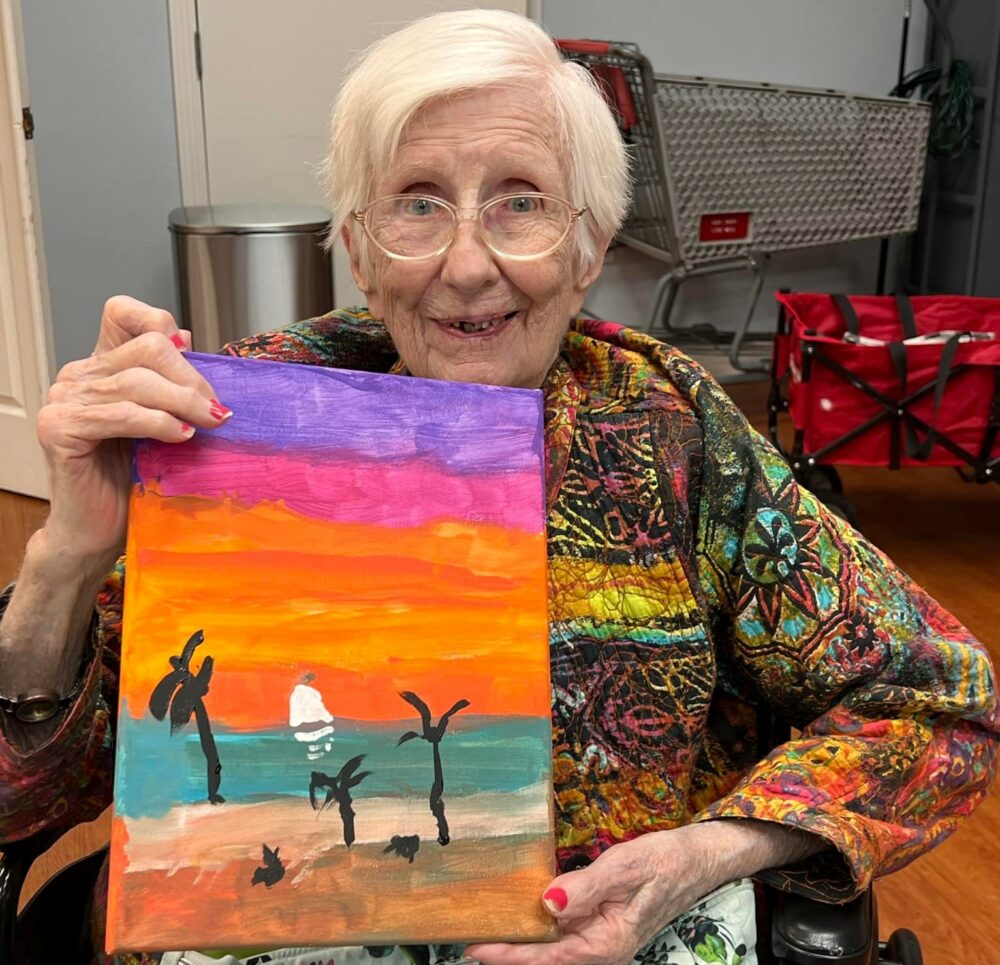 senior woman smiles and holds her completed painting of a beach