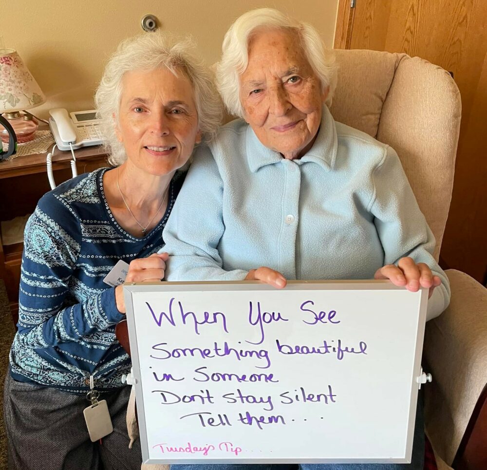 senior woman and woman caregiver pose together with a sign