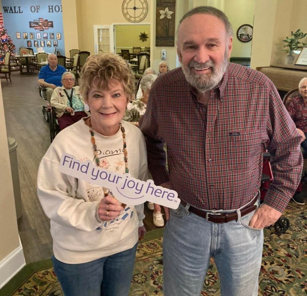 senior man and senior woman smile together while holding a sign that says "find your joy here"