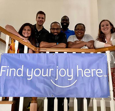 six employees smile with a "find your joy here" sign on the staircase