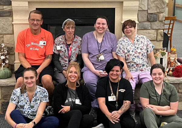eight senior living employees smile together sitting by a fireplace